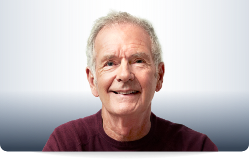 Ein 64 Jahre alter Herr mit grauem Haar und einem lila Pullover.